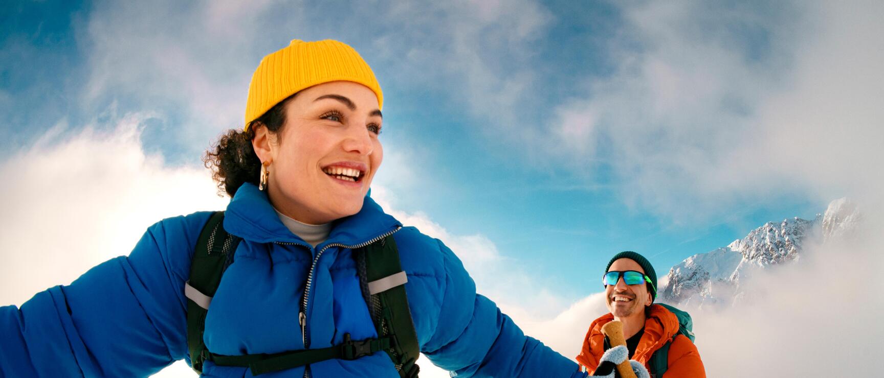 Zwei Schneeschuhwandernde bewegen sich lachend durch die verschneite Berglandschaft der österreichischen Alpen. Die kräftigen Farben der Winterkleidung, die weichen Wolken und die weite Schneekulisse vermitteln Lebensfreude, Leichtigkeit und das gemeinsame Erleben eines aktiven Wintertags in der Natur.