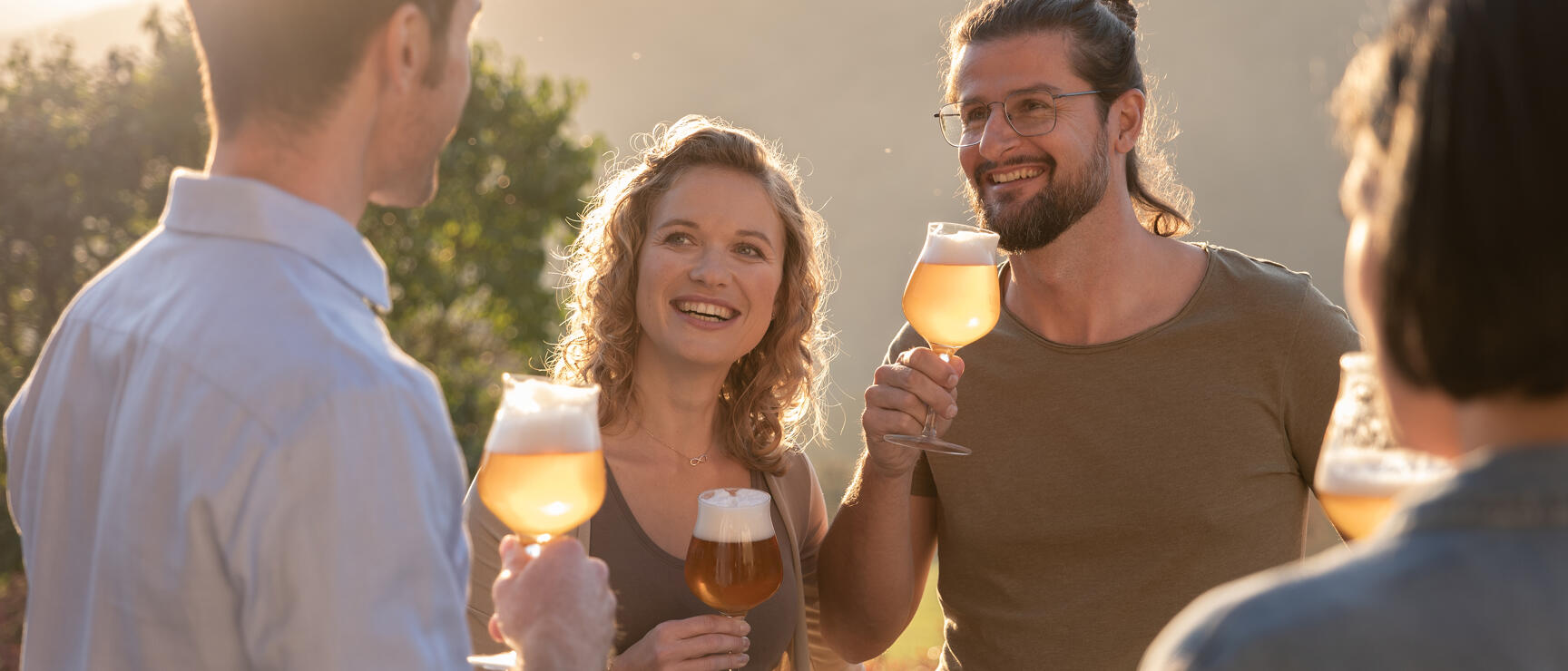 Four people with beer glasses outdoors at sunset, hills in the background.