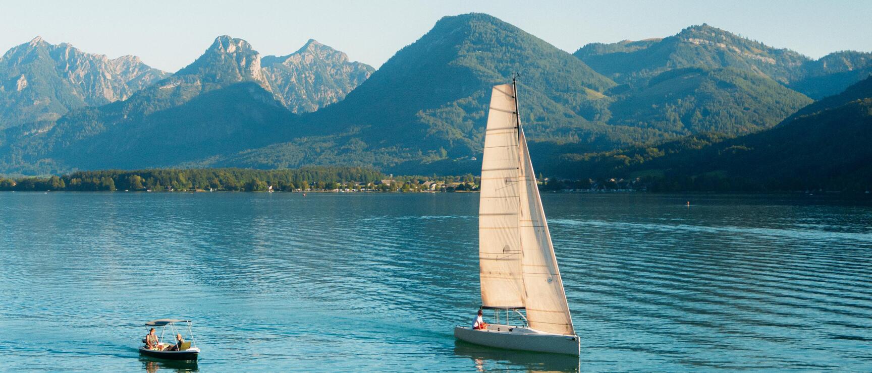 Segelboot und Motorboot auf einem ruhigen See, grüne Berge im Hintergrund, blauer Himmel.