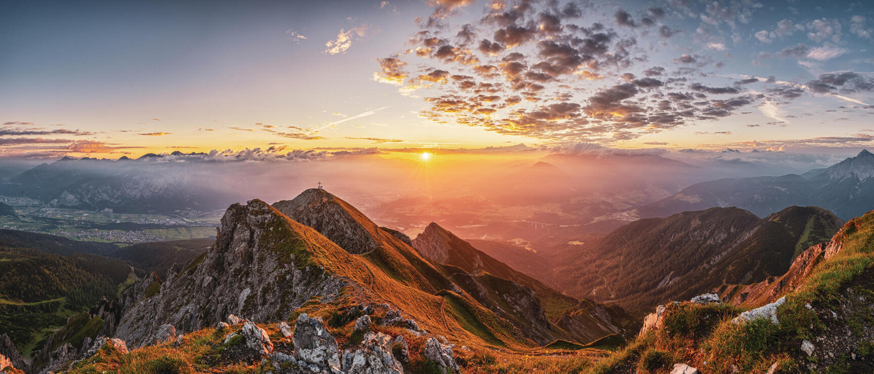 Rocky mountain ridge at sunset, valley with city in background, summit cross visible on neighbouring peak