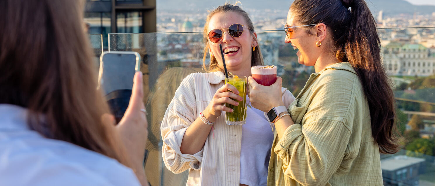 Zwei Frauen mit Cocktails auf einer Dachterrasse, Wien-Panorama im Hintergrund, dritte Person fotografiert sie.