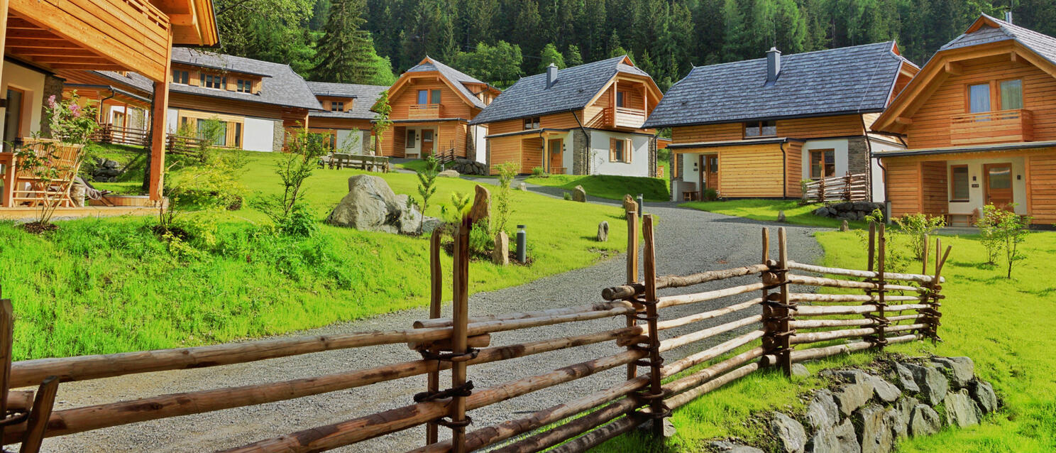Several wooden chalets on green meadow, gravel path with wooden fence, forested mountain in background.
