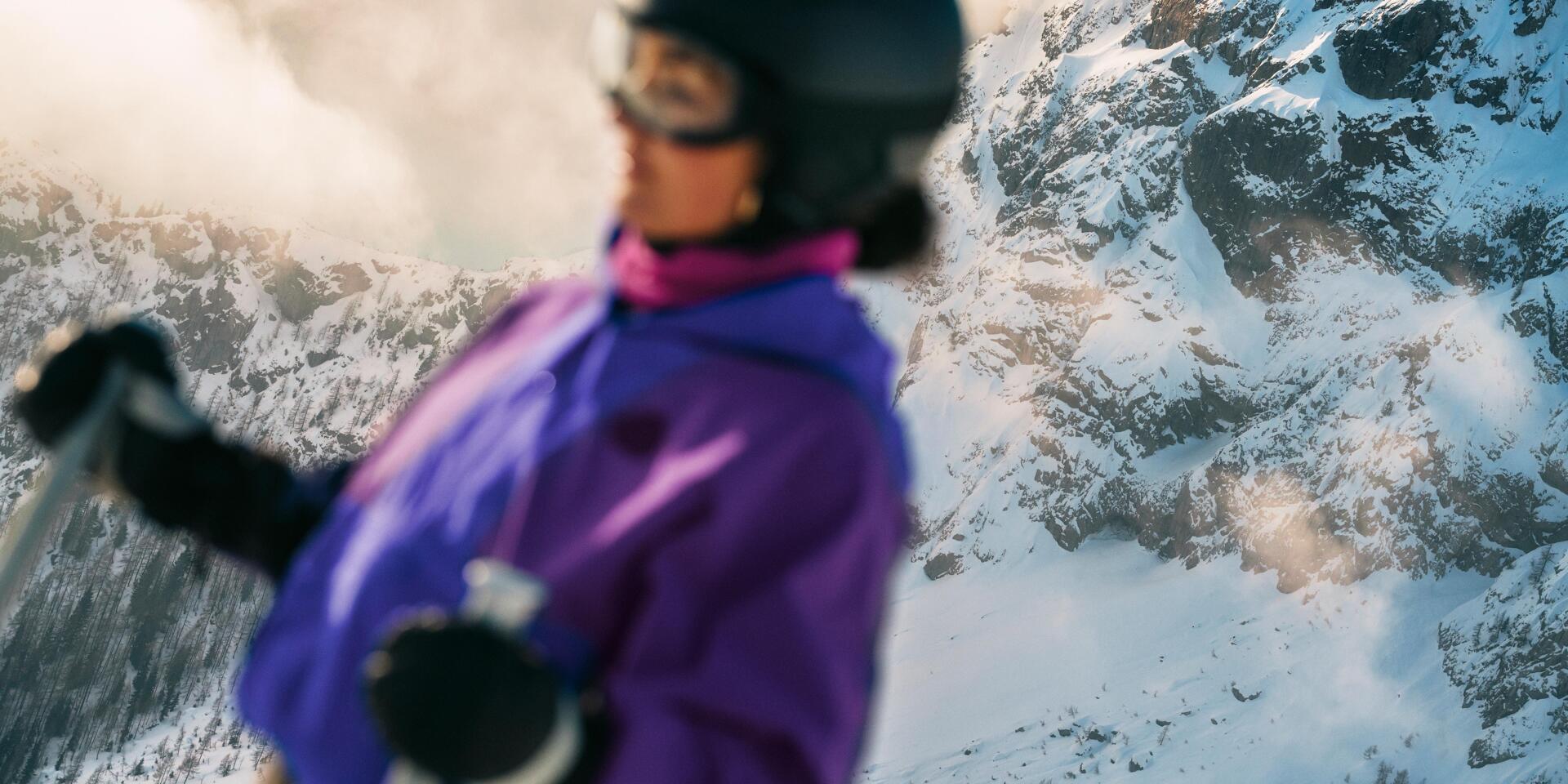 Im Vordergrund ist eine Skifahrerin in violetter Winterbekleidung zu sehen, während im Hintergrund schneebedeckte Felsberge die eindrucksvolle alpine Winterlandschaft prägen.
