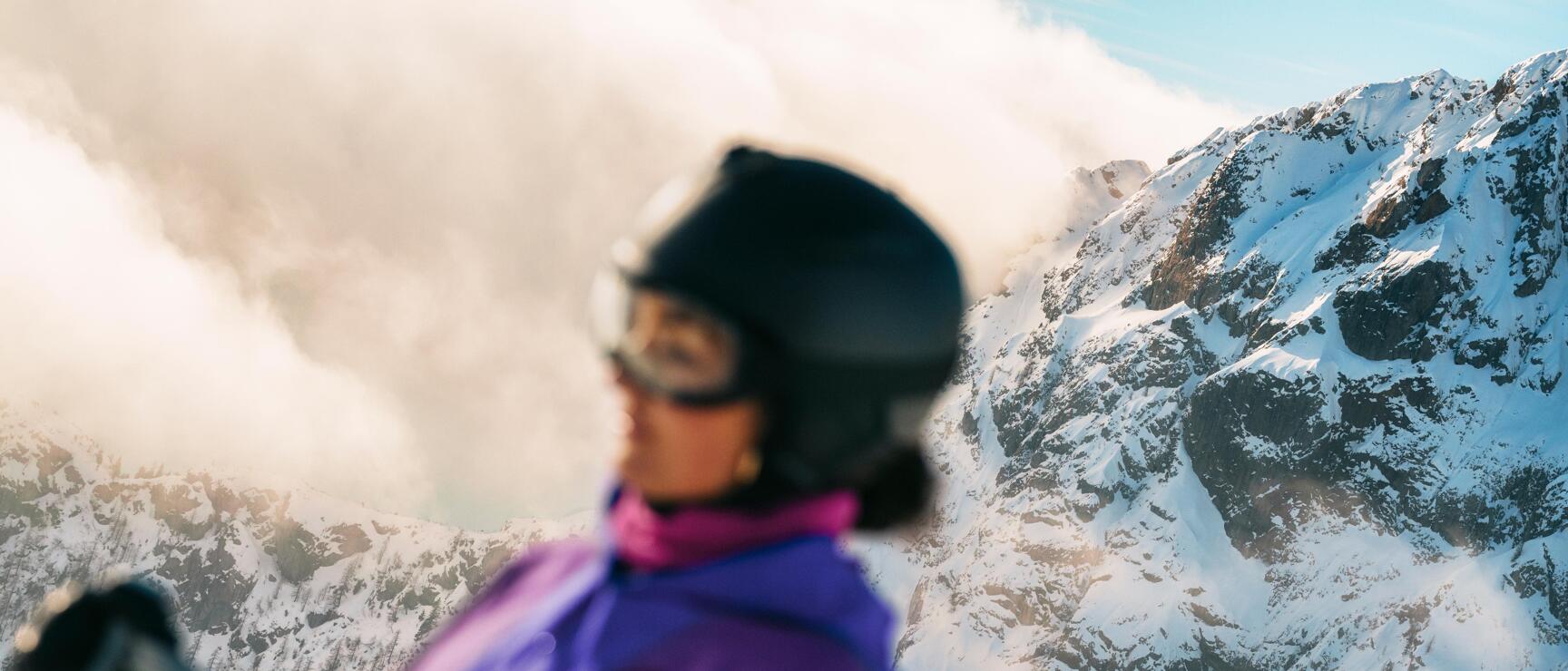 Im Vordergrund ist eine Skifahrerin in violetter Winterbekleidung zu sehen, während im Hintergrund schneebedeckte Felsberge die eindrucksvolle alpine Winterlandschaft prägen.