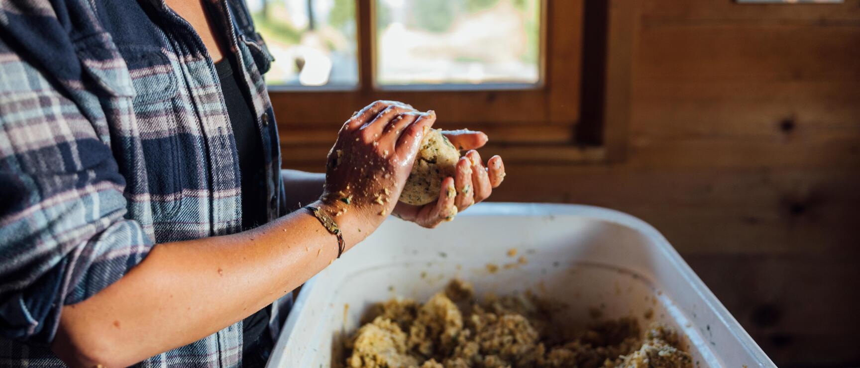 Woman shaping a dumpling with hands from dough in white bowl, wooden room in background.