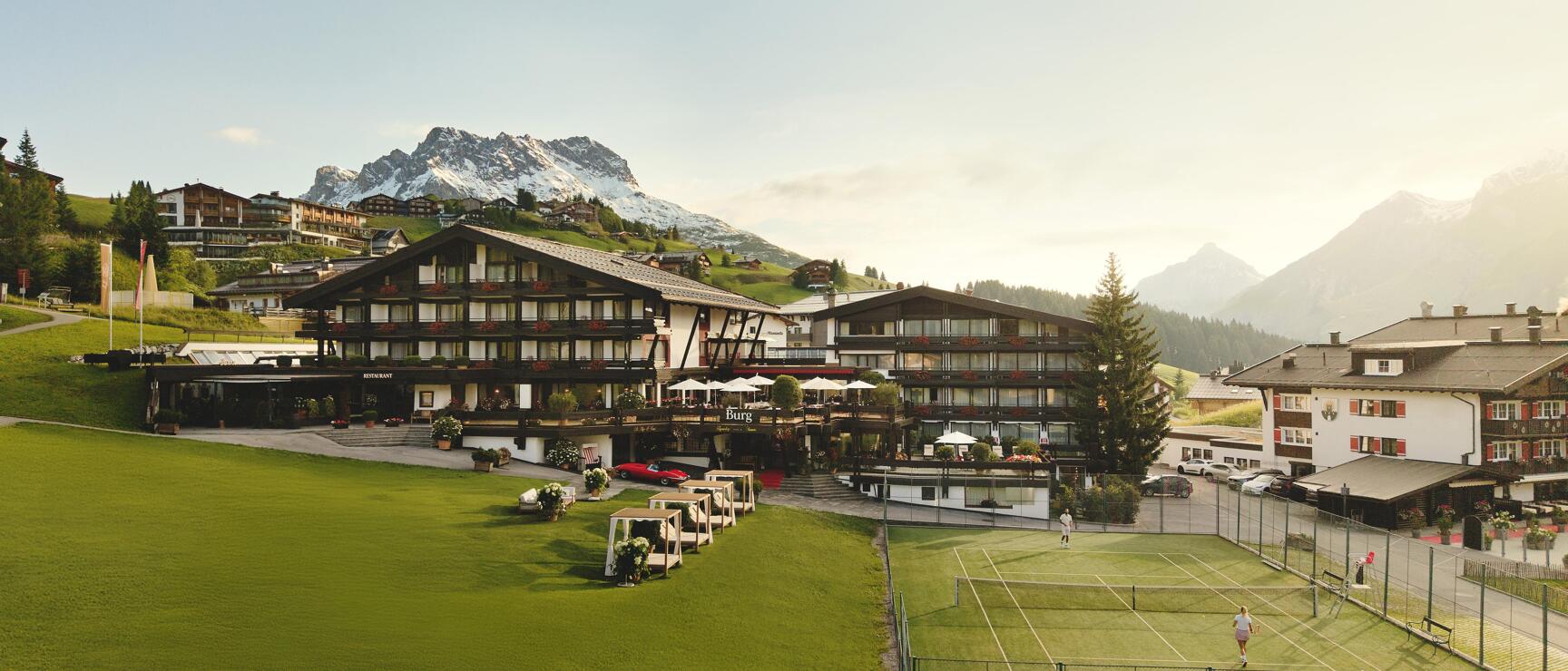 Hotel Burg in Lech am Arlberg with tennis court, green meadow and snow-capped peak in the background.