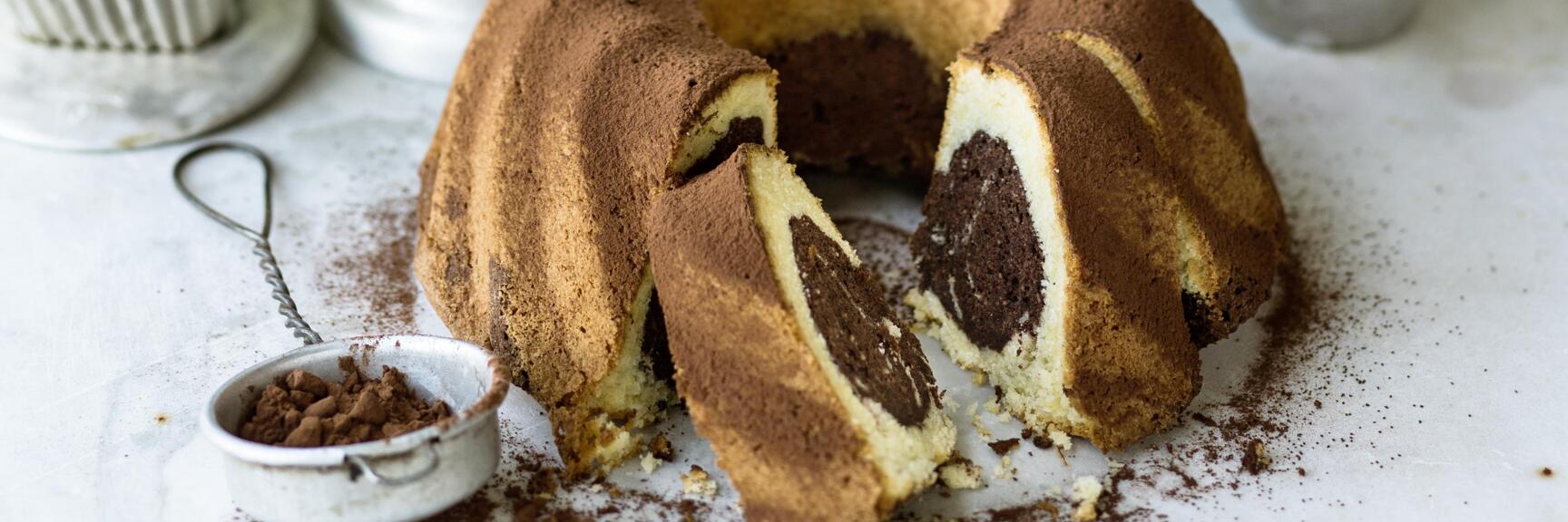 Angeschnittener Marmorgugelhupf mit Kakaopulver bestäubt, daneben Backutensilien auf hellem Untergrund.