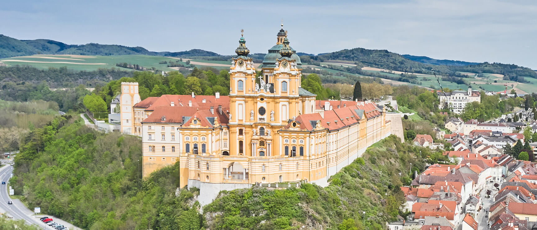 Aerial view of Melk Abbey on a rocky cliff, yellow Baroque facade, red roofs, village and hills in background.