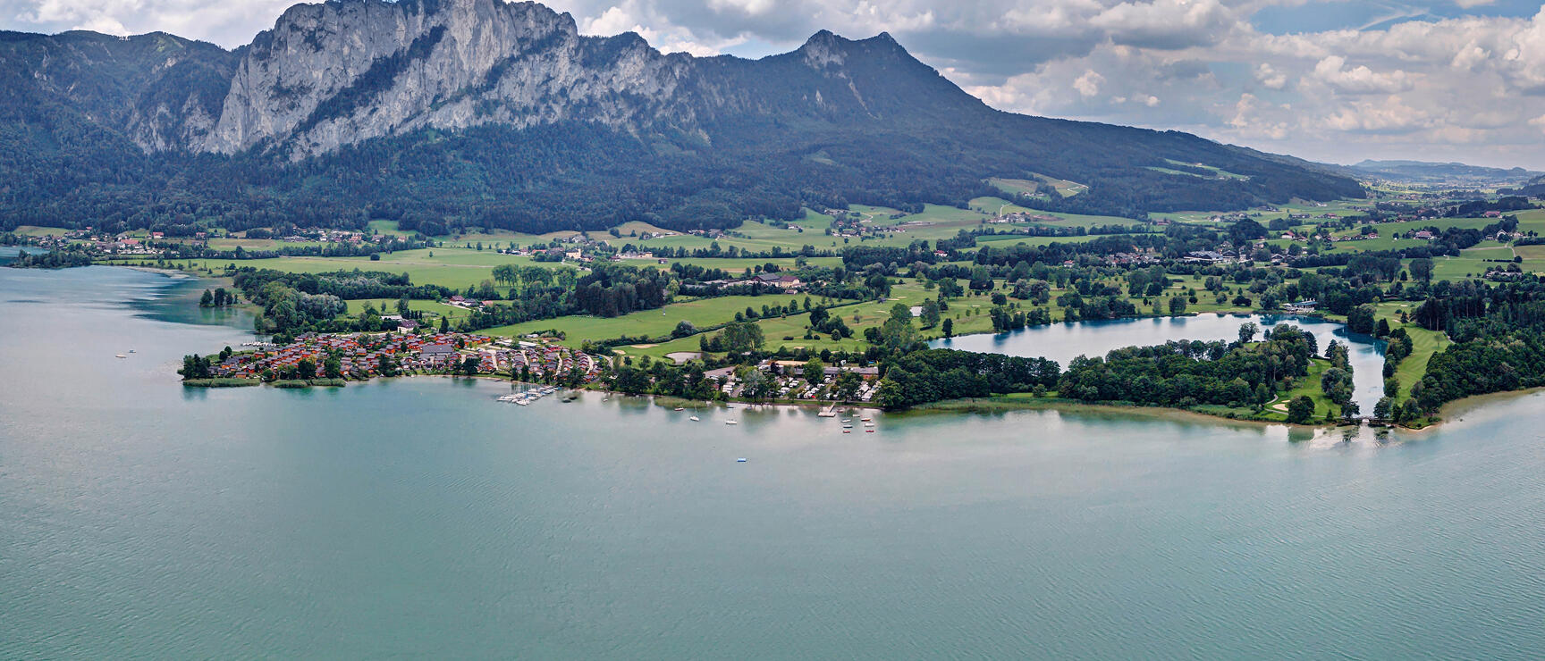 Luftaufnahme der Gemeinde St. Lorenz: Blick vom Mondsee auf den Ort, im Hintergrund die Drachenwand und ein wolkiger Himmel.