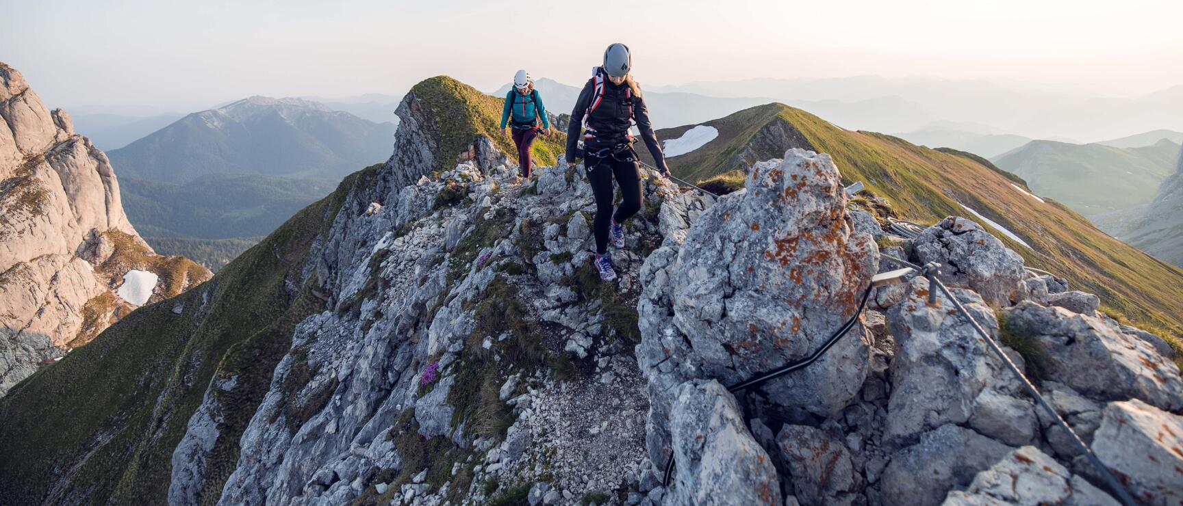 Two people with helmets and backpacks on a rocky ridge, climbing rope, wide mountain panorama.