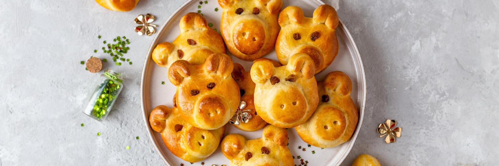 Goldbraune Hefeteig-Brötchen in Schweinchenform auf einem Teller, mit goldenen Kleeblatt-Deko und grünem Streugut.