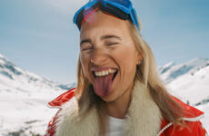 Woman with blue ski goggles and red jacket sticking out her tongue, snow-covered mountains in the background.