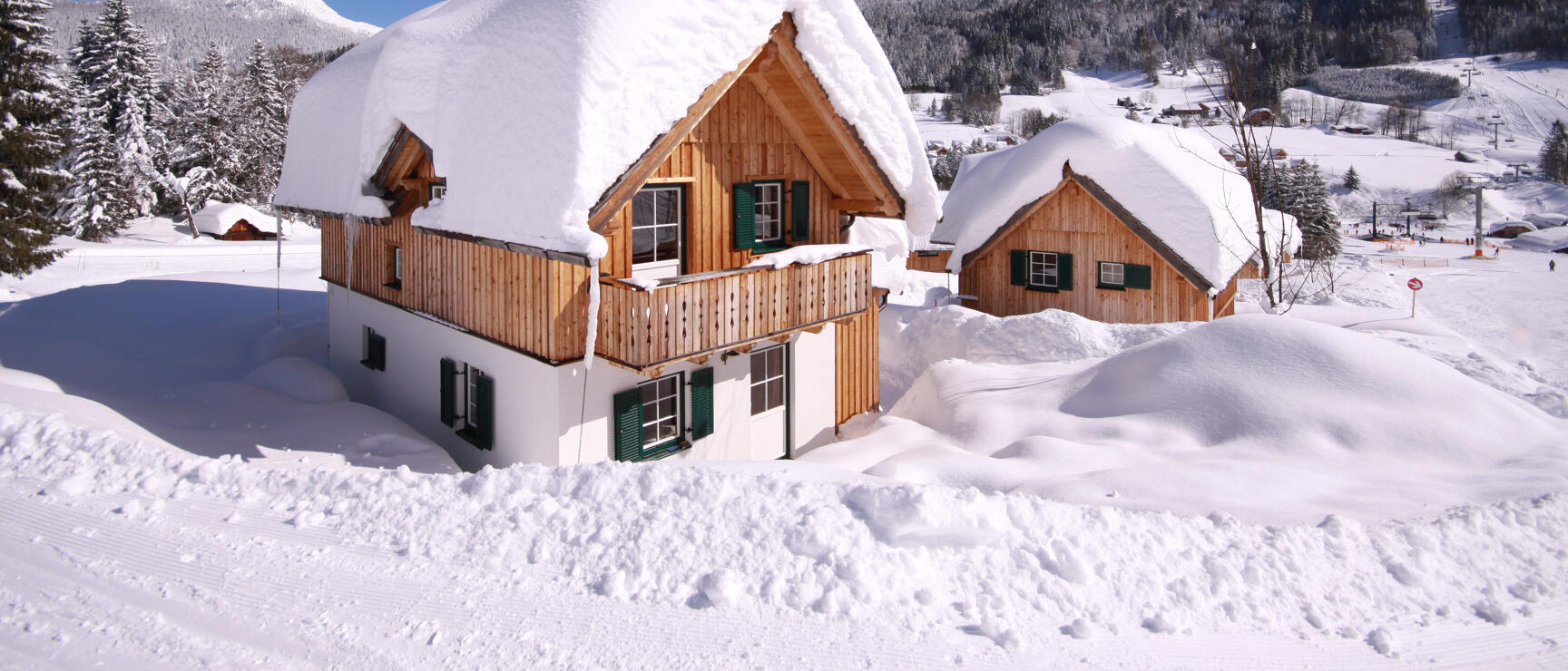 Snow-covered wooden chalets with green shutters, mountain panorama and ski area in the background under blue sky.