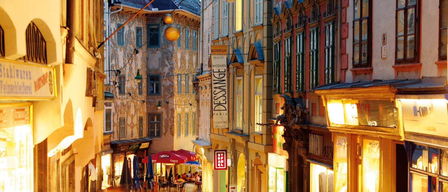 Busy shopping street in Graz with illuminated shops, pedestrians and ornate facade in background.