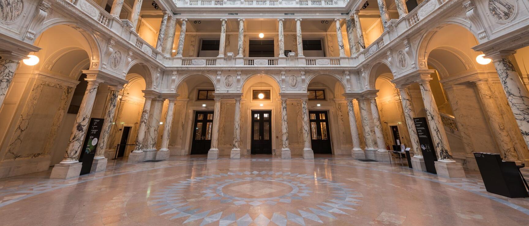 Grand hall with marble columns, frescoed ceiling and glass roof, two galleries stacked, marble floor.