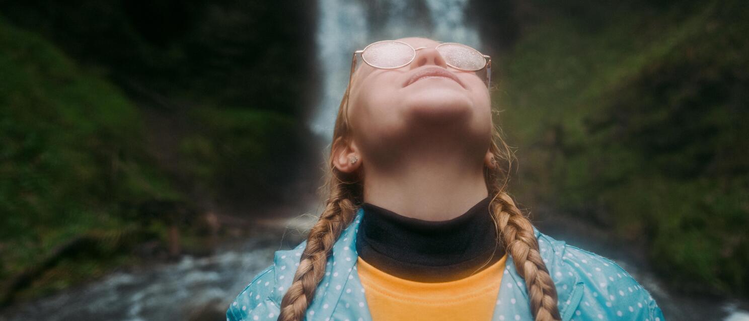 Woman with braids and sunglasses looking upward, waterfall with green rocks in background.
