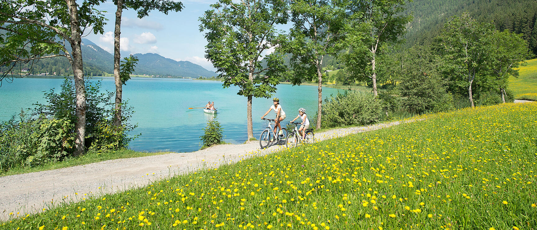 Two female cyclists wearing helmets on a gravel path beside a turquoise lake, mountain panorama in the background.