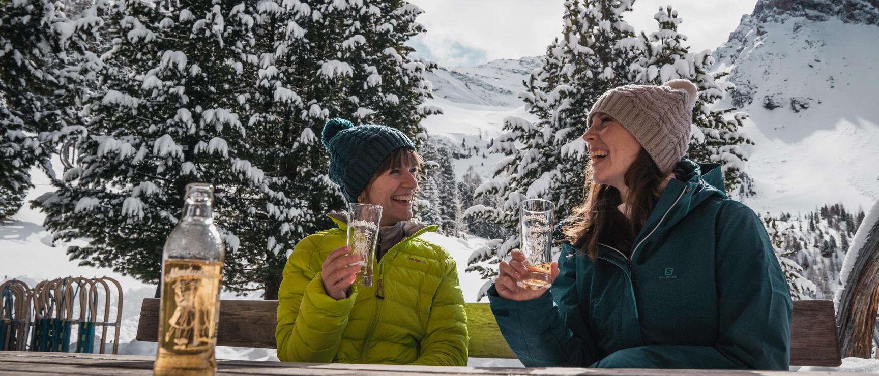 Two women with drinks at a wooden table on an alpine hut terrace, snow-covered mountains in the background.