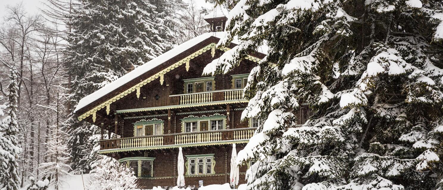 Snow-covered wooden chalet with green balconies and ornaments, surrounded by snow-laden fir trees.