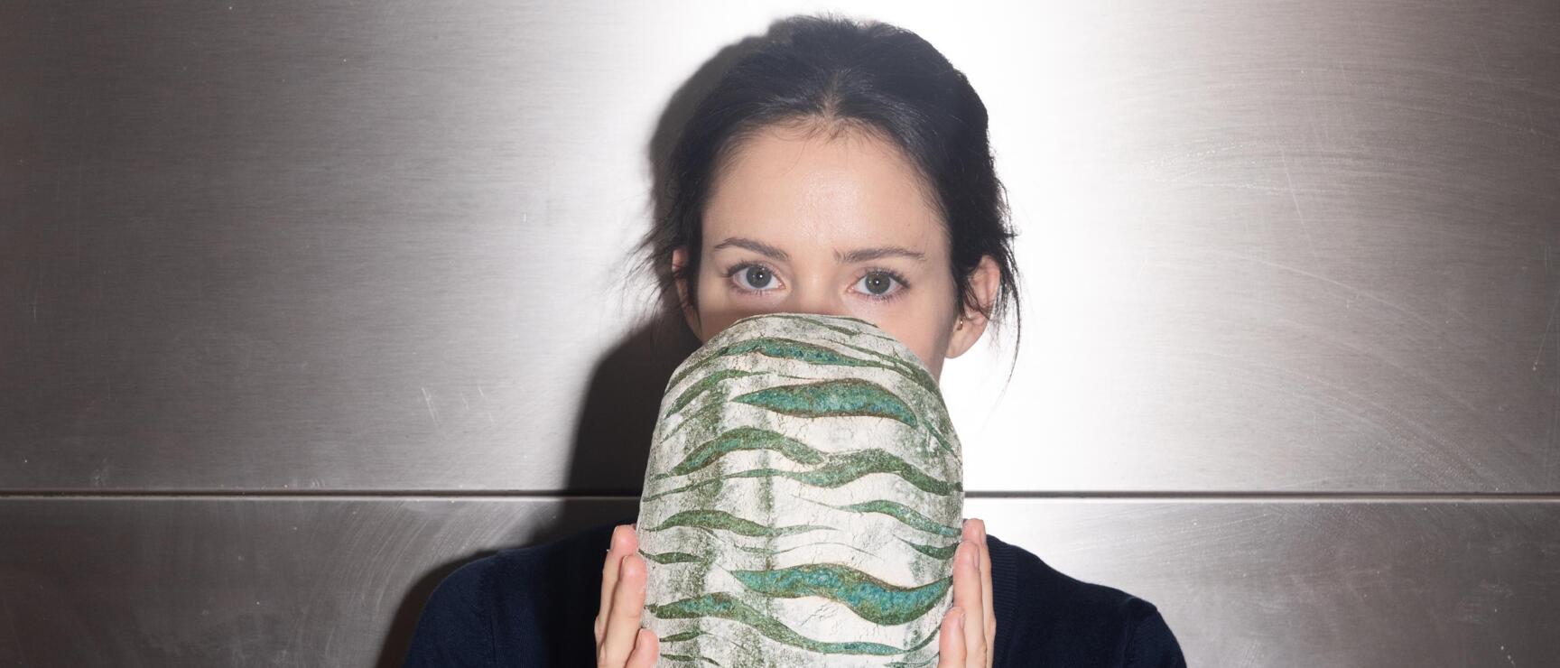 Frau hält ovales Brot mit grün-weißem Muster vor ihr Gesicht, metallische Wand im Hintergrund.