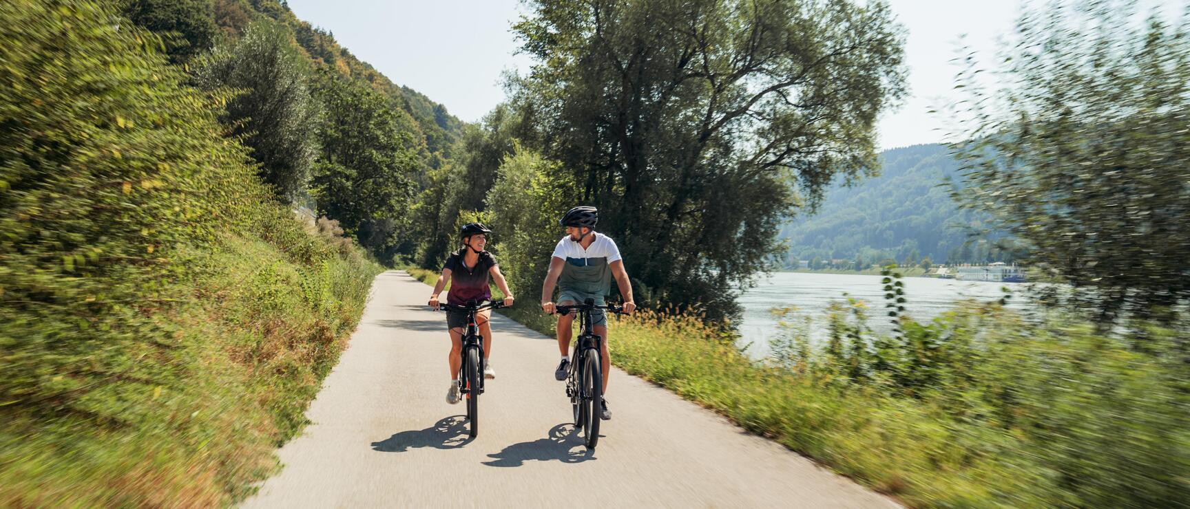 Two cyclists wearing helmets on a path along the Danube with forested hills in the background.