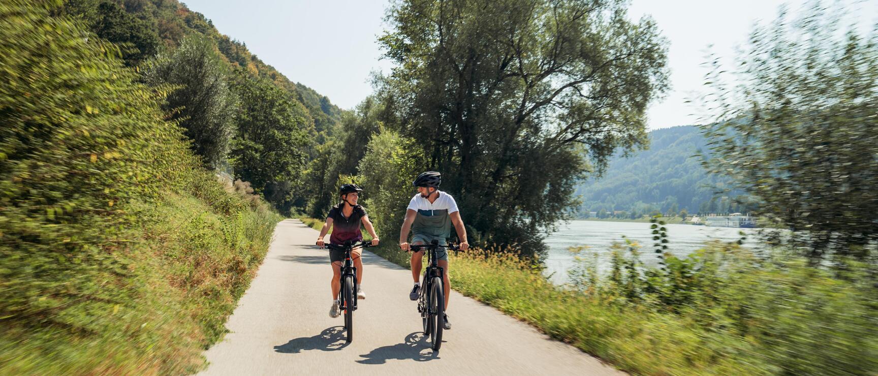 Two cyclists wearing helmets on a path along the Danube with forested hills in the background.