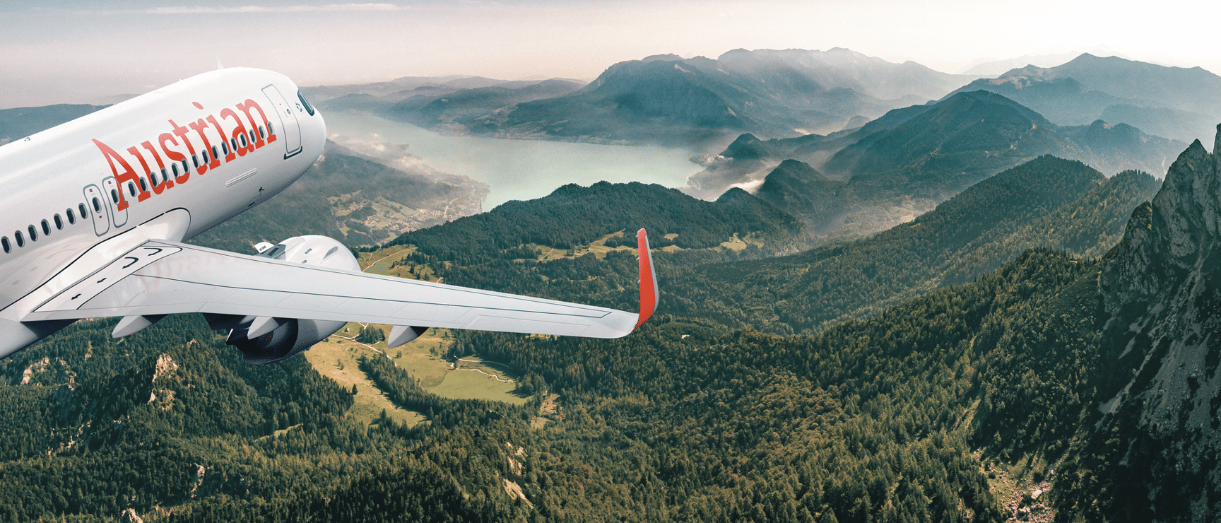 Austrian Airlines aircraft flying over forested mountains and a lake, mountain range in background.