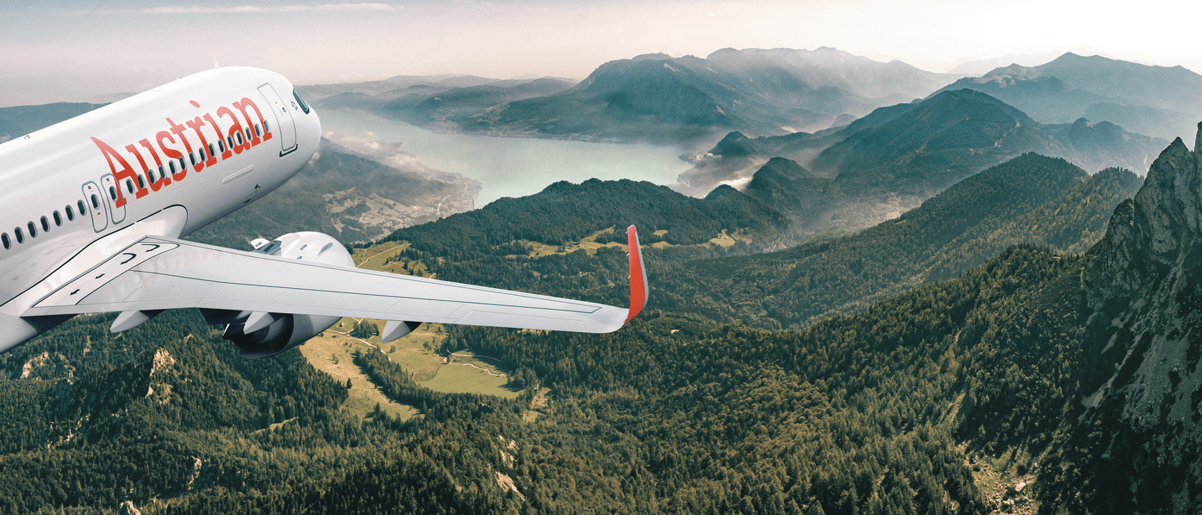 Austrian Airlines aircraft flying over forested mountains and a lake, mountain range in background.