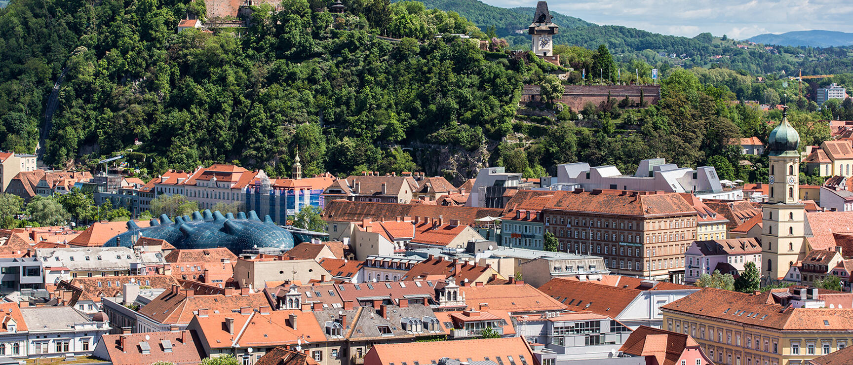 Panoramic view of Graz with red tile roofs, Schlossberg with clock tower and art house roof in foreground.