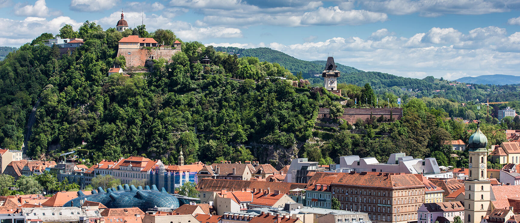 Panoramic view of Graz with red tile roofs, Schlossberg with clock tower and art house roof in foreground.