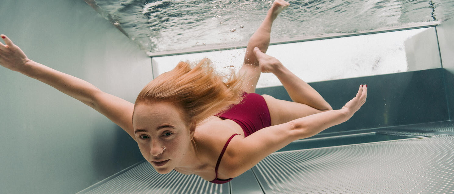 Unterwasseraufnahme: Frau mit roten Haaren im roten Bikini schwimmt in einem Pool, Metallboden sichtbar.