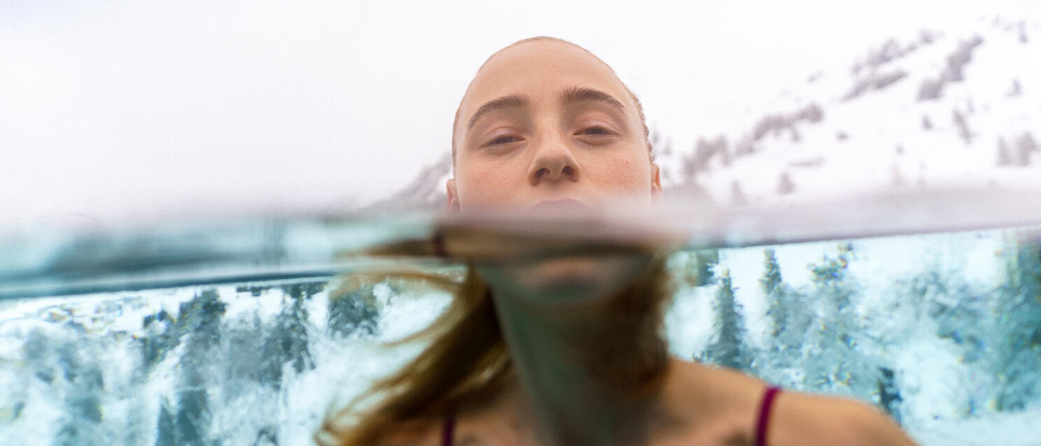 Woman in outdoor pool with face at water surface, snow-capped mountains and pine trees in background.