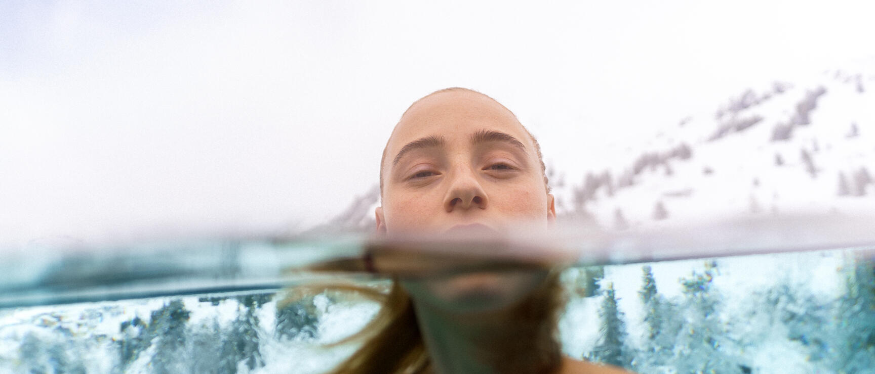 Woman in outdoor pool with face at water surface, snow-capped mountains and pine trees in background.
