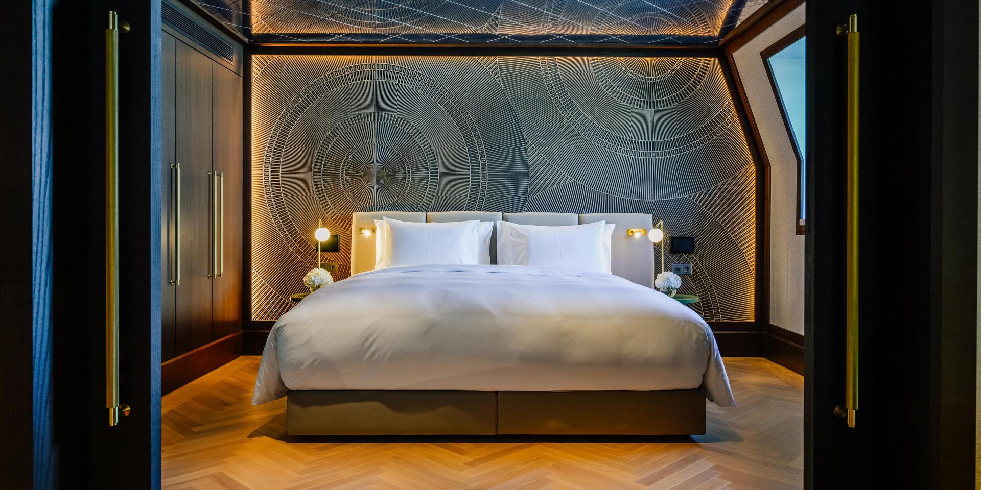 Hotel room with double bed, white bedding, patterned wall, wooden floor and gold door handles.