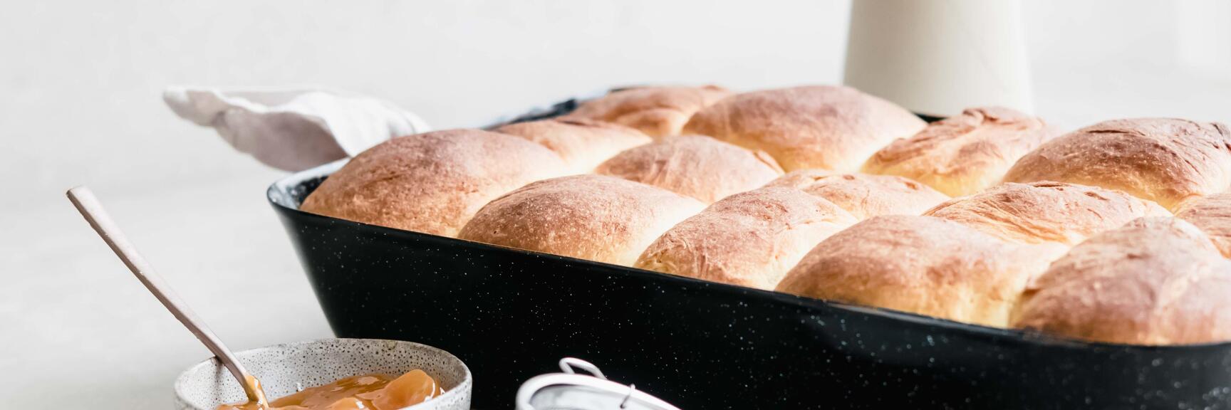 Freshly baked buchteln in black baking dish, next to bowl of jam and sieve with icing sugar.