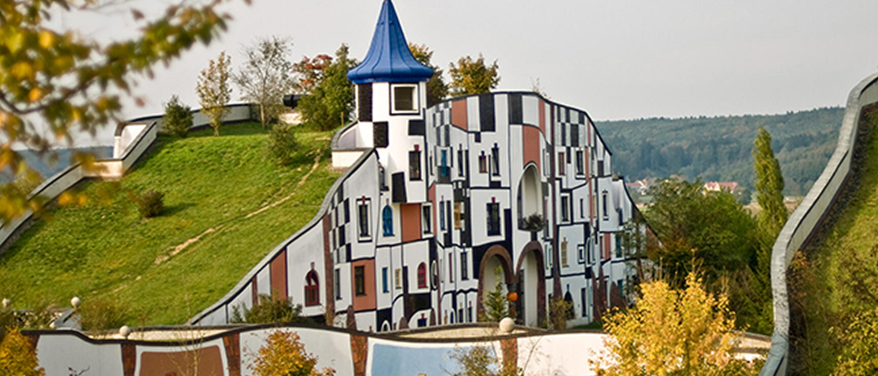 Hundertwasser building with colorful checkered façade and blue tower, green roof, trees in foreground.