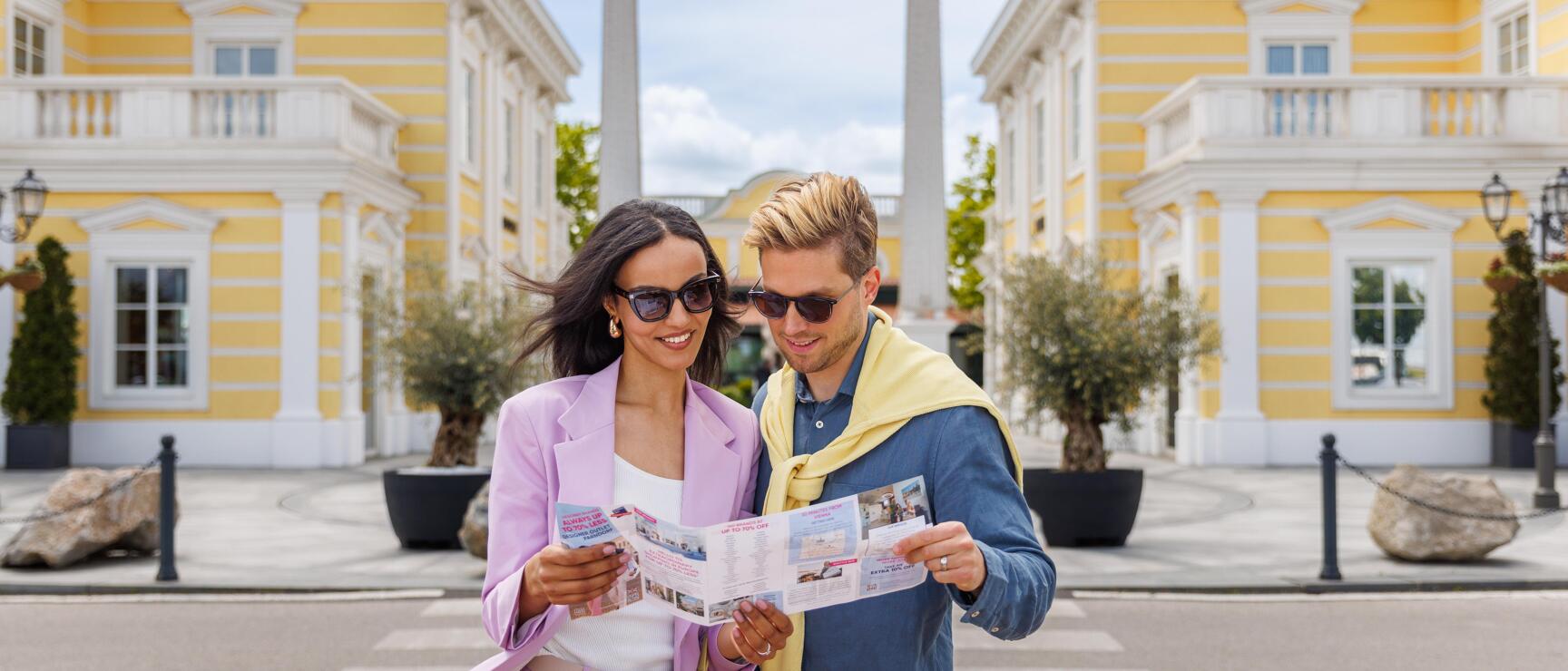 Two people wearing sunglasses reading a flyer in front of a yellow building with obelisks.