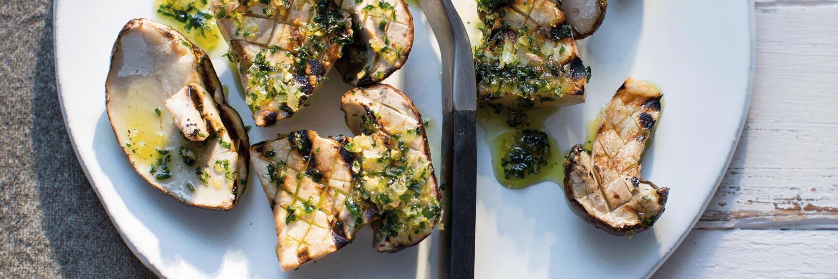Grilled porcini mushrooms with herb oil sauce on white plate, black fork, light wooden background.