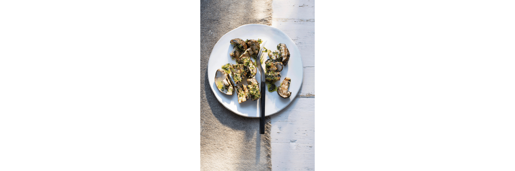 Grilled porcini mushrooms with herb oil sauce on white plate, black fork, light wooden background.