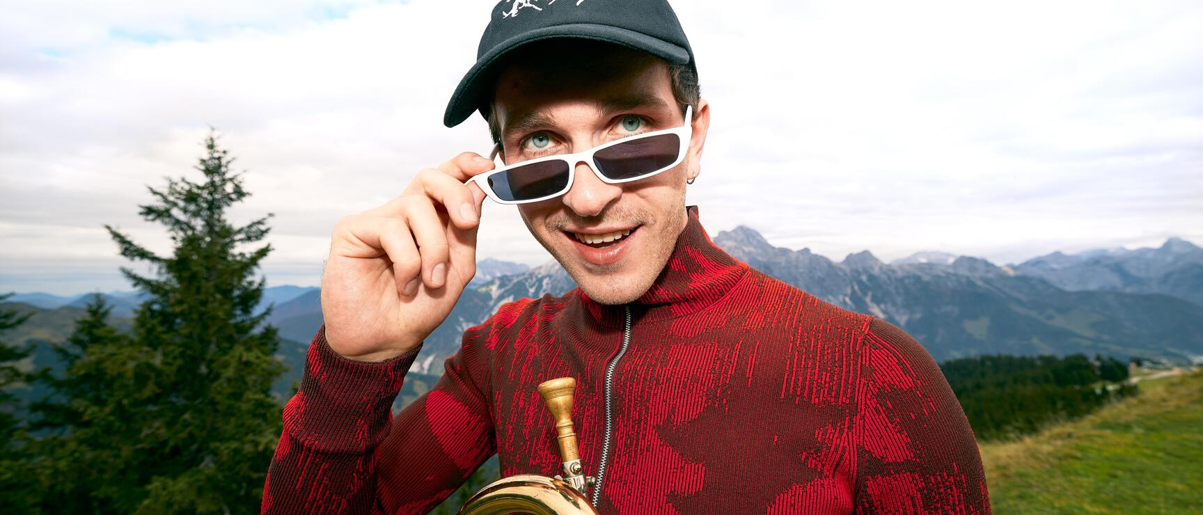 Man with trumpet, white sunglasses and red sweater on a mountain peak, alpine panorama in the background.