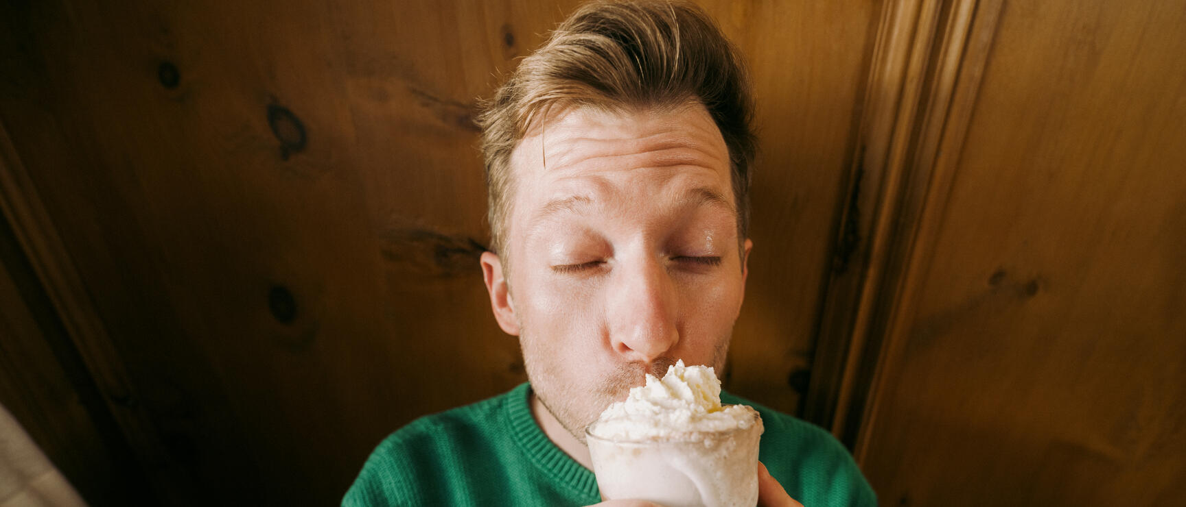 Man in green sweater drinking coffee with whipped cream from a glass, wooden panelling in background.