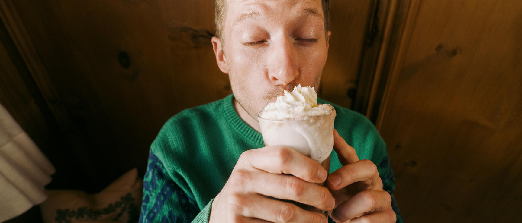 Man in green sweater drinking coffee with whipped cream from a glass, wooden panelling in background.