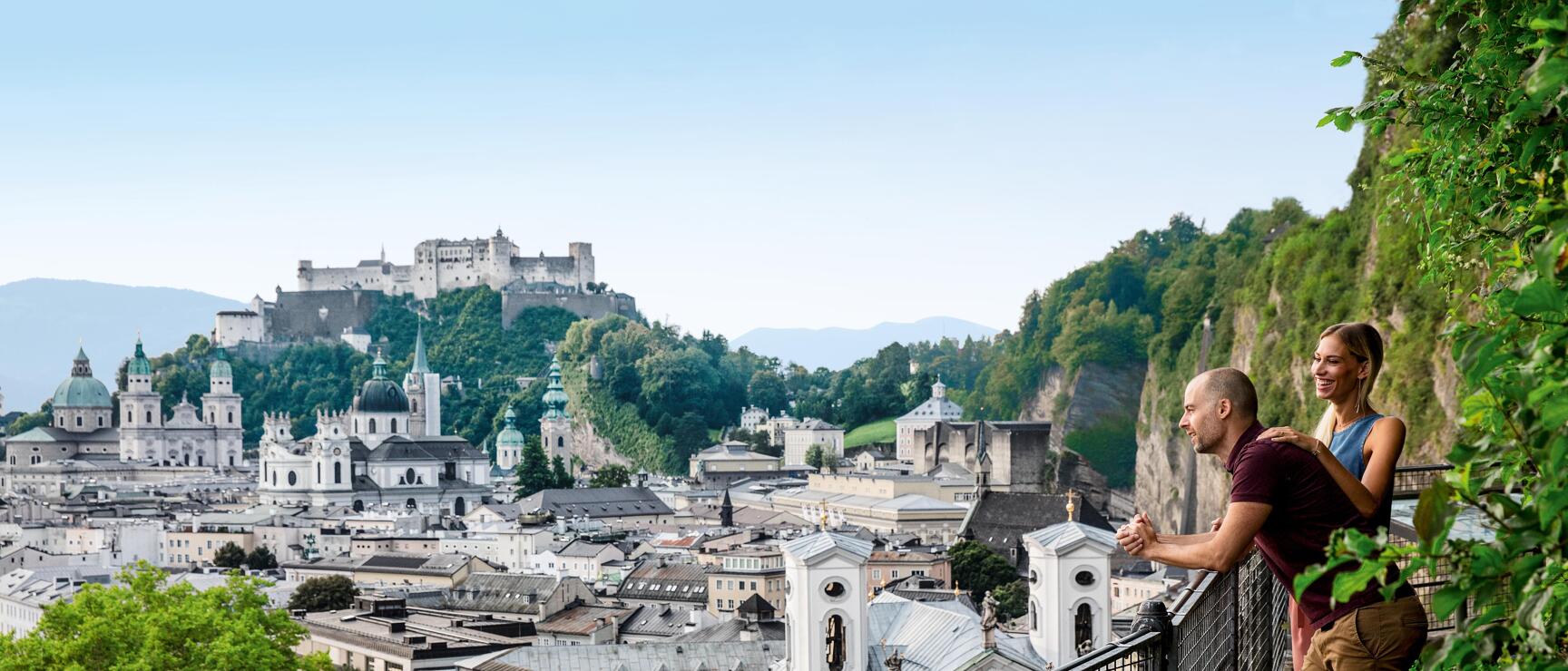 Two people at a viewpoint overlooking Salzburg's old town and Hohensalzburg Fortress.
