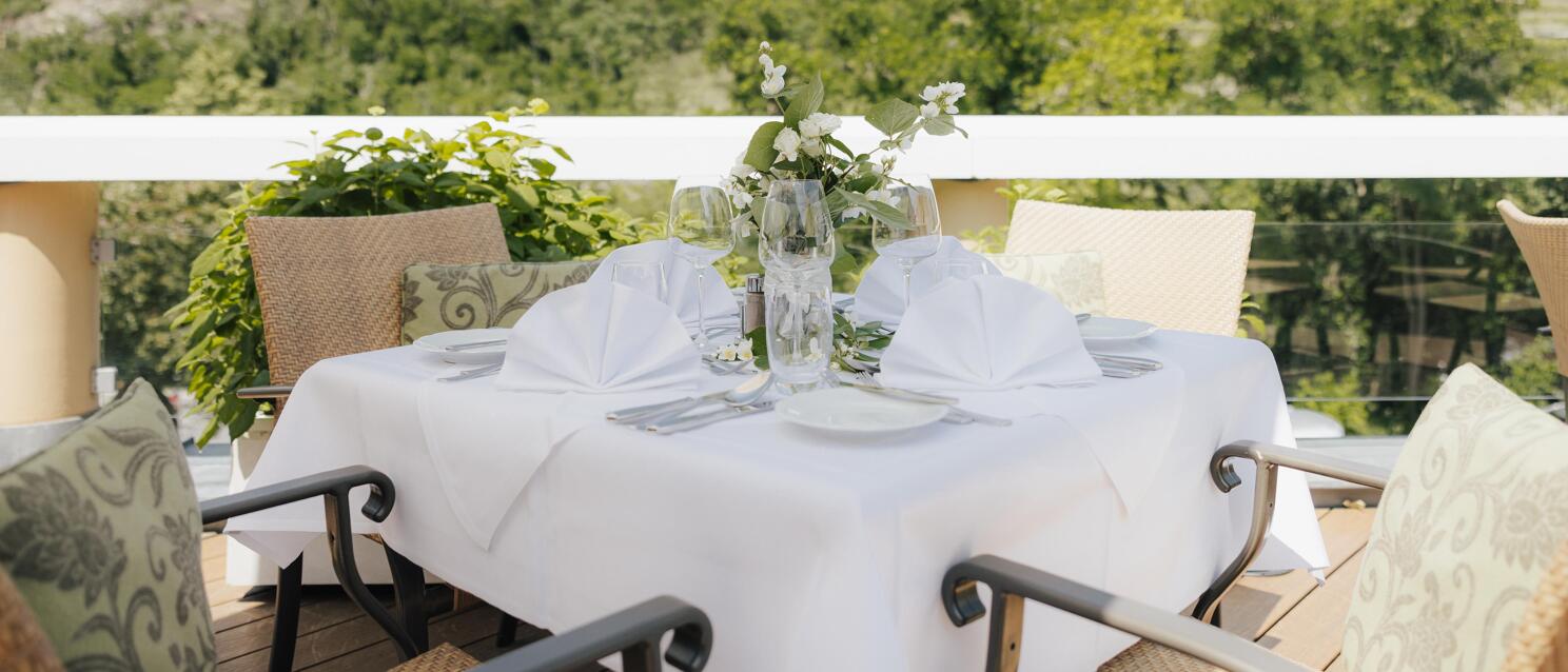 Laid table with white tablecloth and flower bouquet on a terrace, vineyard hills in the background.