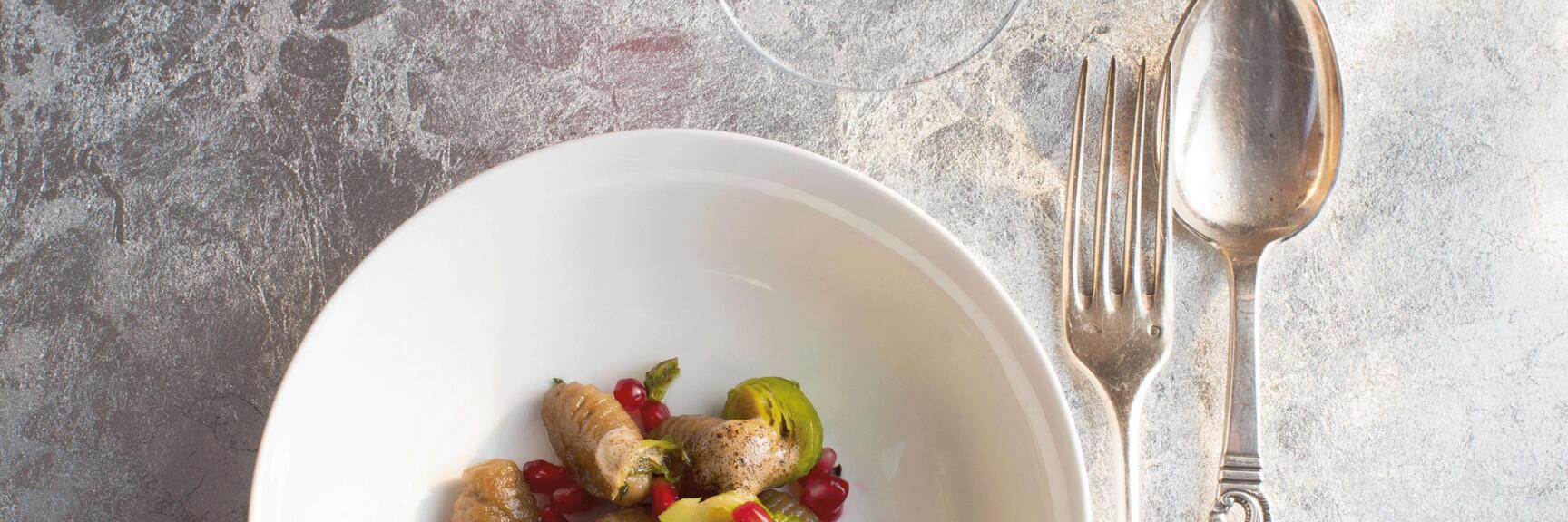 Gnocchi with Brussels sprouts and pomegranate seeds in white bowl, red wine and water glass, silver cutlery.