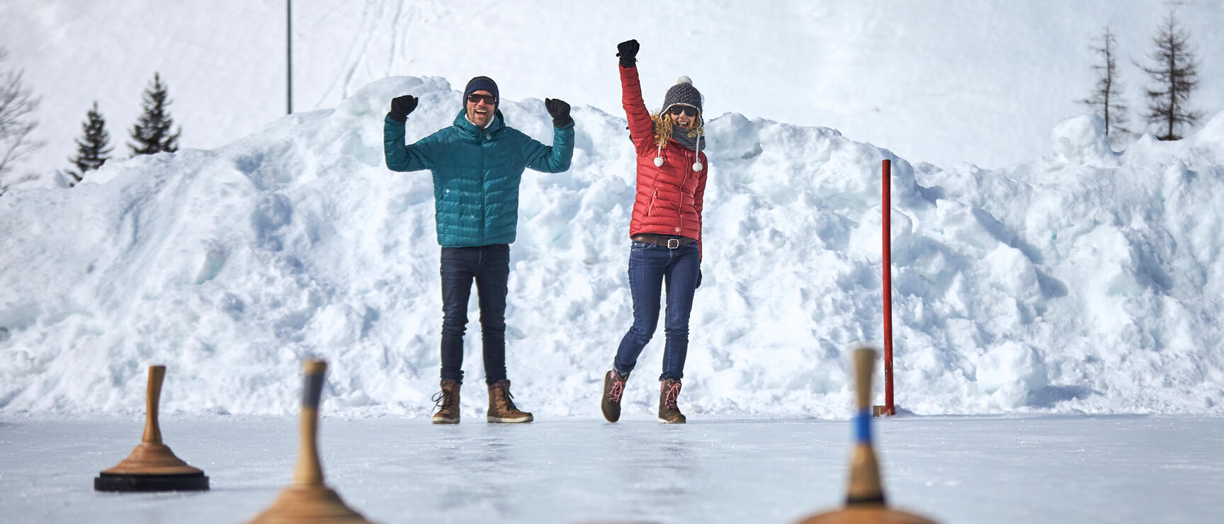 Zwei Personen jubeln auf einer Eisstockbahn mit Eisstöcken im Vordergrund, Schneehaufen im Hintergrund.