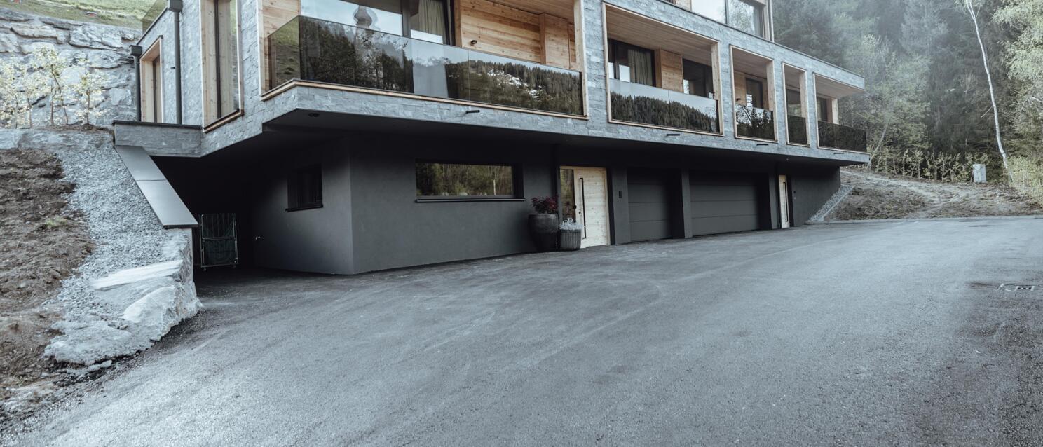 Modern chalet with wood and stone façade, glass balcony, garage in basement, forest in background.