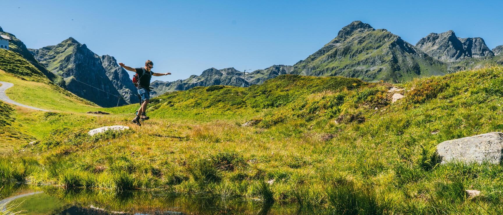 Person jumping over an alpine pond, water reflection, green mountain meadows and rocky peaks in the background.