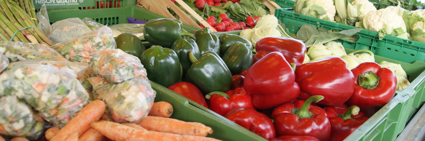 Vegetable stand with carrots, red and green peppers, radishes, tomatoes and cauliflower in green crates.