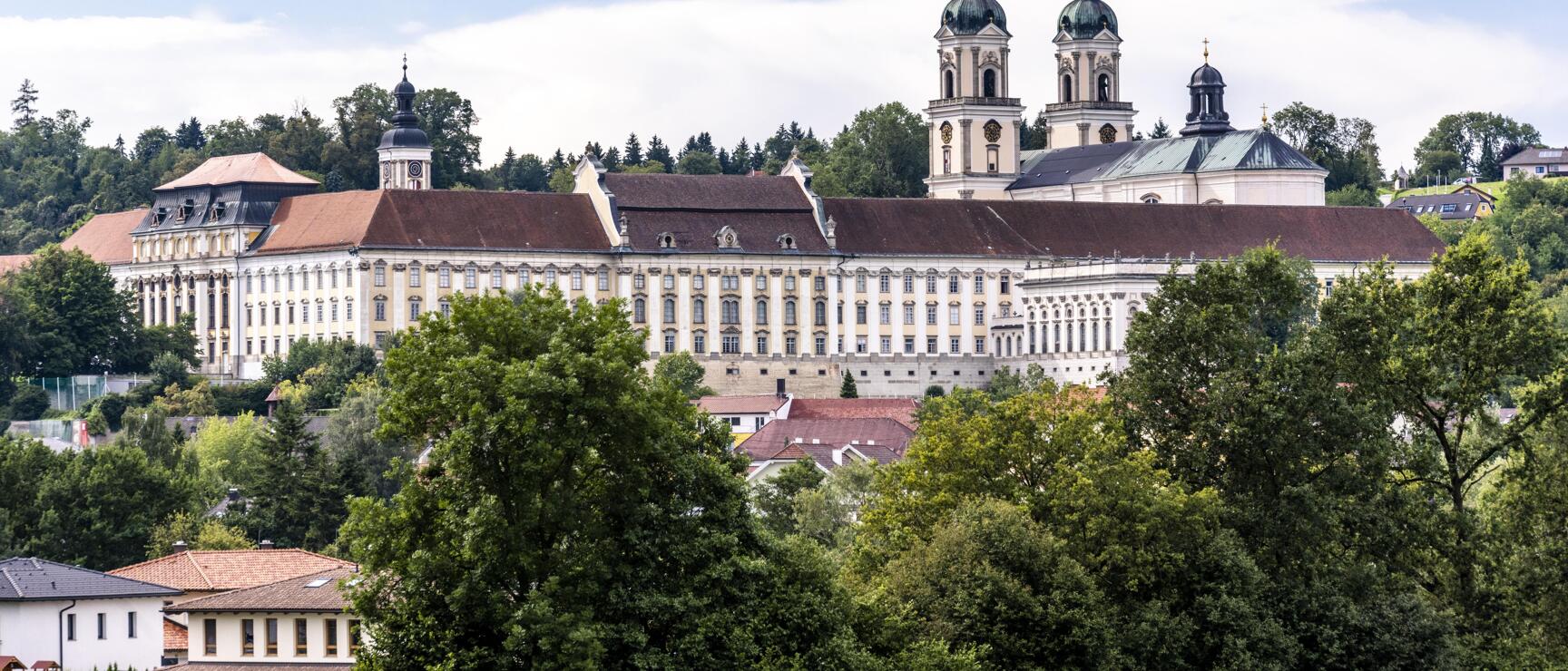 Stift Kremsmünster with two church towers and long Baroque facade, surrounded by trees, blue sky.